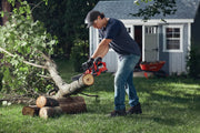 Photo of a person using a CRAFTSMAN chainsaw, SKU CMCCS610B, cutting a tree branch in a yard with a shed in the background.