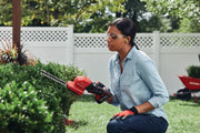 Person using Craftsman CMCSS800C1 cordless shrub shear to trim bushes in a garden, wearing gloves and ear protection.