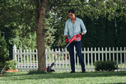 Photo of a person using a Craftsman string trimmer CMCST915B trimming grass in a yard near a tree and white fence.