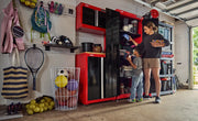 CRAFTSMAN red and black storage cabinets are shown in a garage, used for organizing sports equipment and other items. Two people, an adult and a child, are sorting items in the cabinets. The scene also includes shelves, hooks, and bins with sports gear like balls, rackets, and backpacks.