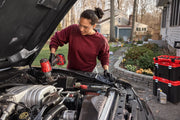 A person with a blurred face is working on a car engine outdoors, using a red cordless power tool. They are wearing black